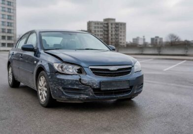 Uszkodzony niebieski samochód osobowy z wgniecionym przednim zderzakiem na parkingu na tle bloków mieszkalnych